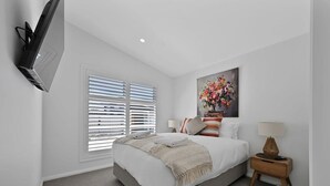 A bedroom with a bed, a TV mounted on the wall, a window with blinds, a painting of flowers, and a bedside table with a lamp.