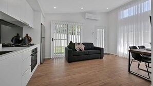 A modern kitchen with white cabinets, a black countertop, and a stove. There is a black sofa with pillows and a dining table with chairs. - The Elm Apartments - The Green Room (Orange)