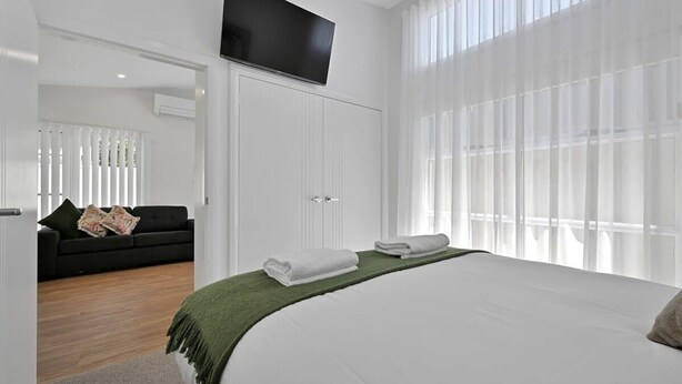 A modern bedroom with a large bed, a TV mounted on the wall, and a view of a balcony. - The Elm Apartments - The Green Room (Orange)