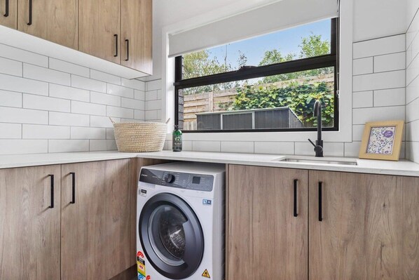 A laundry area with a washing machine, wooden cabinets, a window with a view of greenery, and a tiled backsplash.