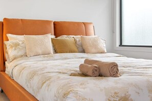 Cama com colcha estampada, várias almofadas e uma toalha enrolada.