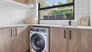 Eine moderne Küche mit Holzausstattung, einer integrierten Waschmaschine, einem Fenster mit Blick ins Grüne und einer weißen Fliesenrückwand.