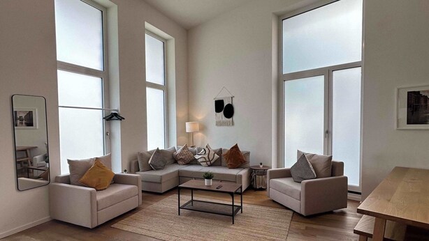 Ein geräumiges Wohnzimmer mit einer großen Eckcouch, einem Couchtisch und einem Esstisch. Große Fenster sorgen für natürliches Licht und einen Blick ins Freie.