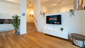 Ein modernes Wohnzimmer mit einem Flachbildfernseher an einer weißen Wand, einer Treppe zu einer weiteren Ebene und einem sichtbaren Küchenbereich im Hintergrund.