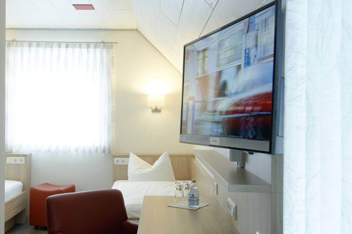 Ein Hotelzimmer mit Bett, Wandfernseher, Schreibtisch und Fernbedienung sowie einem Fenster mit Vorhängen.
