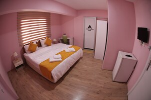 Habitación de hotel en tonos rosados, con cama, mesita de noche, lámpara y armario.