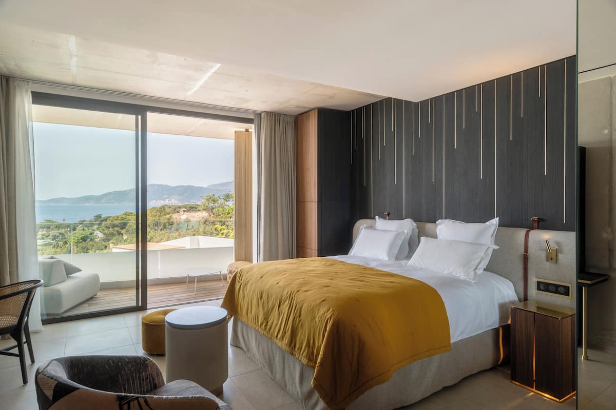 A modern hotel room with a large bed, a sofa, a small round table, and a view of the sea.