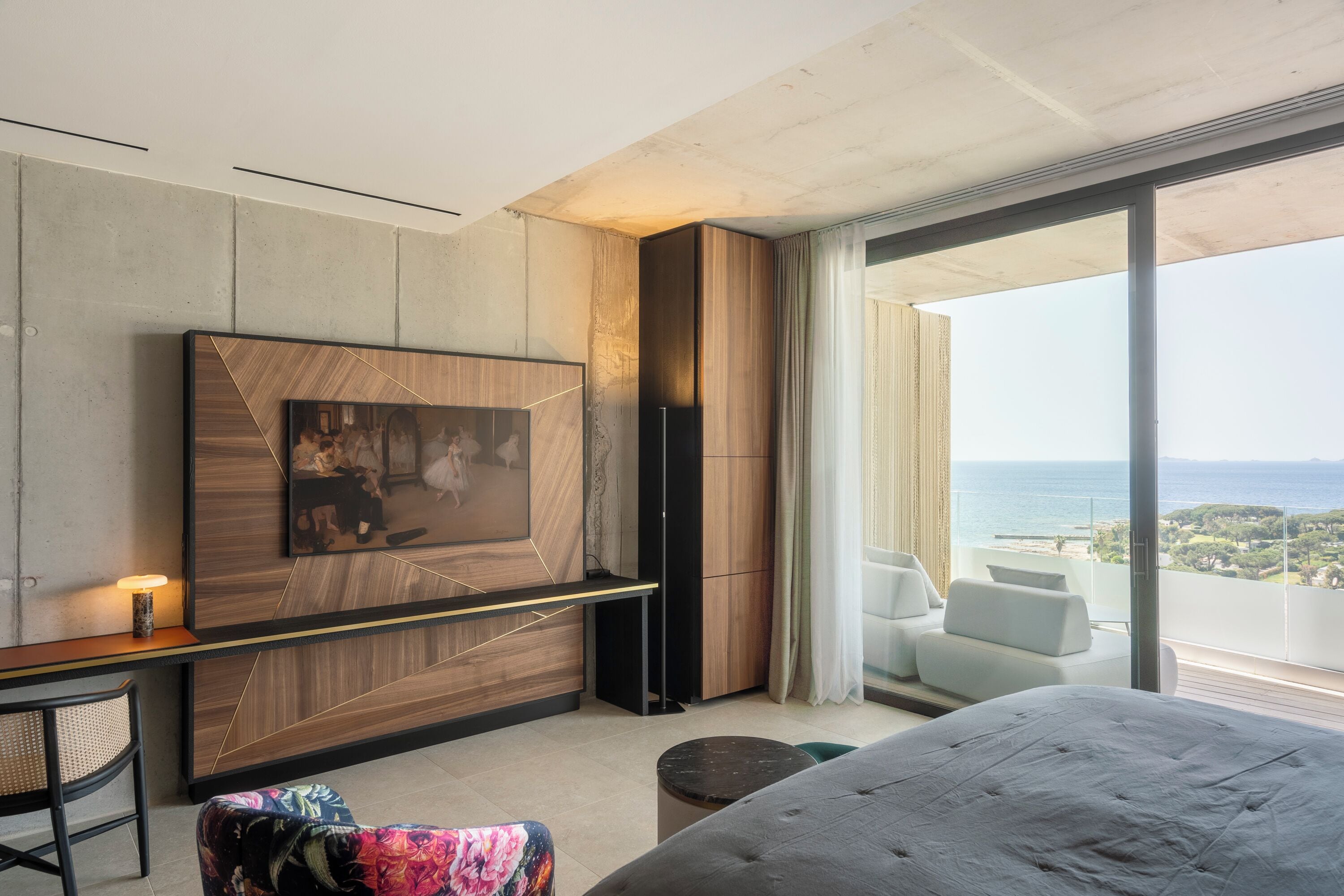 A modern hotel room with a large bed, a desk with a chair, a flat-screen TV mounted on the wall, and a balcony with a view of the sea.