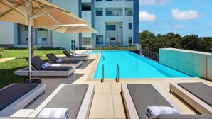 A poolside area with lounge chairs and umbrellas, a swimming pool, and a building in the background. - Luxury Self Catering 1 Bed Apart (Pretoria)