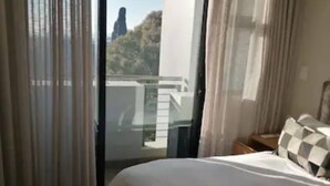 A bedroom with a large bed, a bench, a nightstand, and a view of a building and trees through the window. - Luxury Self Catering 1 Bed Apart (Pretoria)