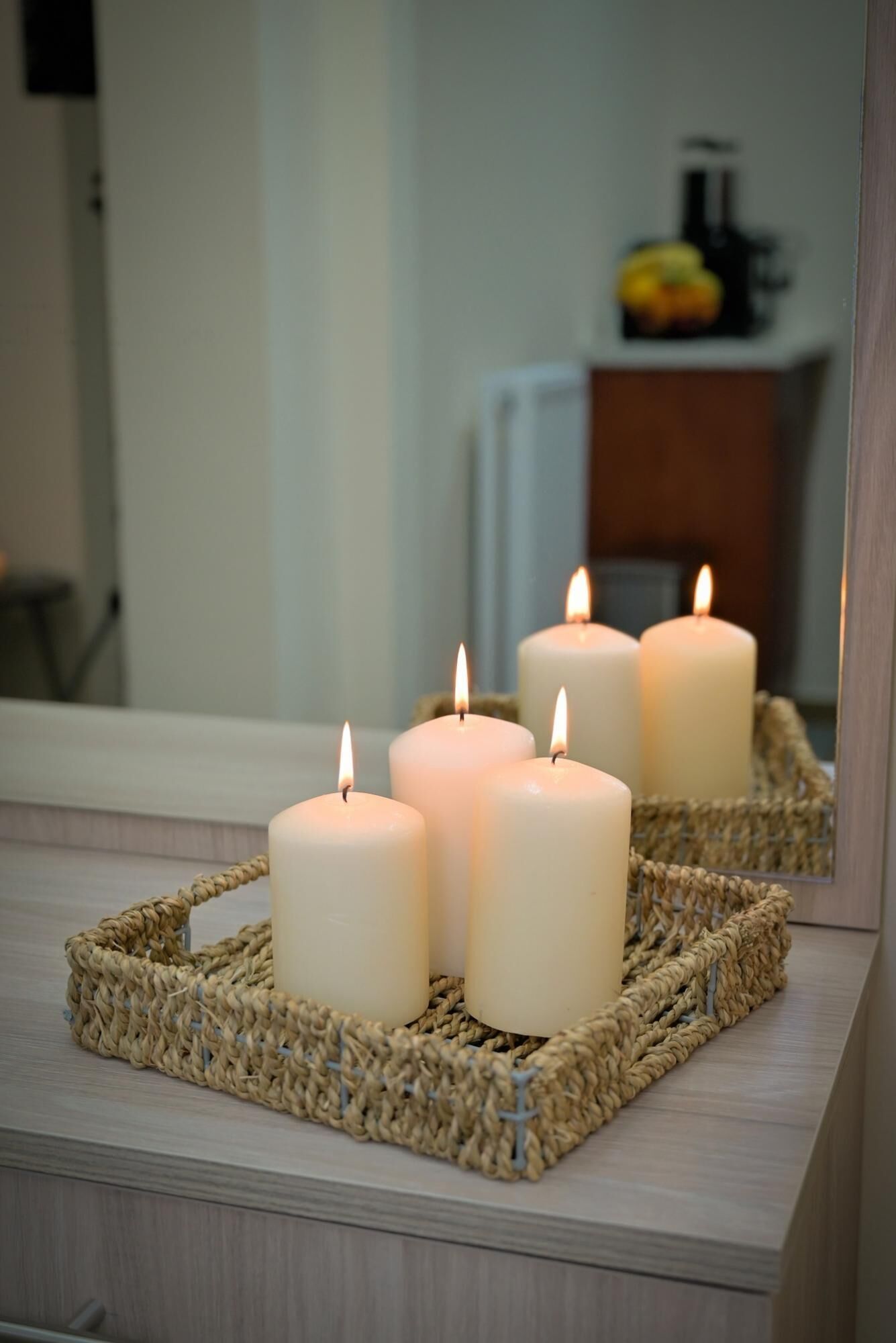 Three lit candles in a woven tray on a wooden surface, with a mirror reflecting a room interior.