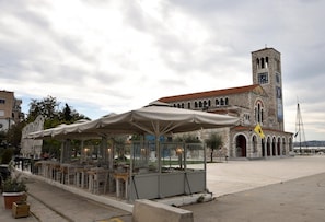 An outdoor café with tables and umbrellas, a stone building with a clock tower, and a clear sky.