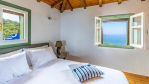 Ein Schlafzimmer mit Holzboden, einem Bett mit weißen Leinentüchern, zwei Fenstern mit grünen Rahmen und Blick aufs Meer.