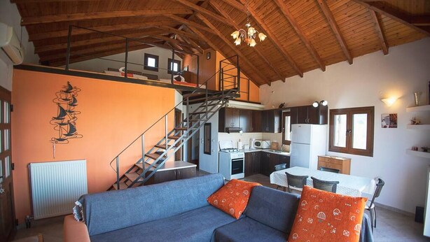 A spacious living area with a blue sofa, orange cushions, a wooden ceiling, a kitchen with modern appliances, and a staircase leading to an upper level.