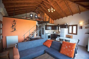 A spacious living area with a blue sofa, orange cushions, a wooden ceiling, a kitchen with modern appliances, and a staircase leading to an upper level. - Foula House in Magnisia (Volos)