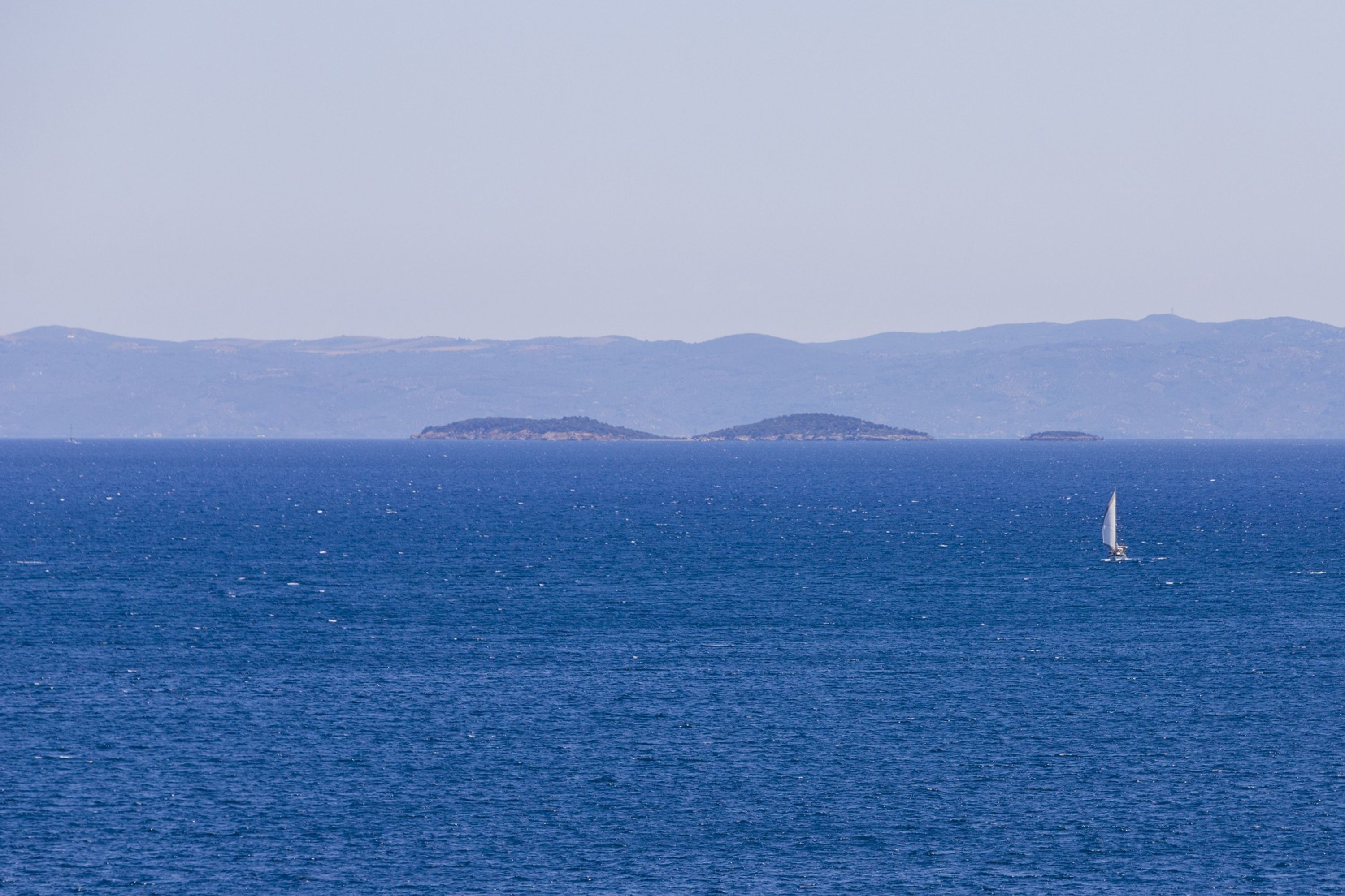 Une vue sur une mer calme, avec un voilier à l’horizon et des reliefs terrestres lointains.