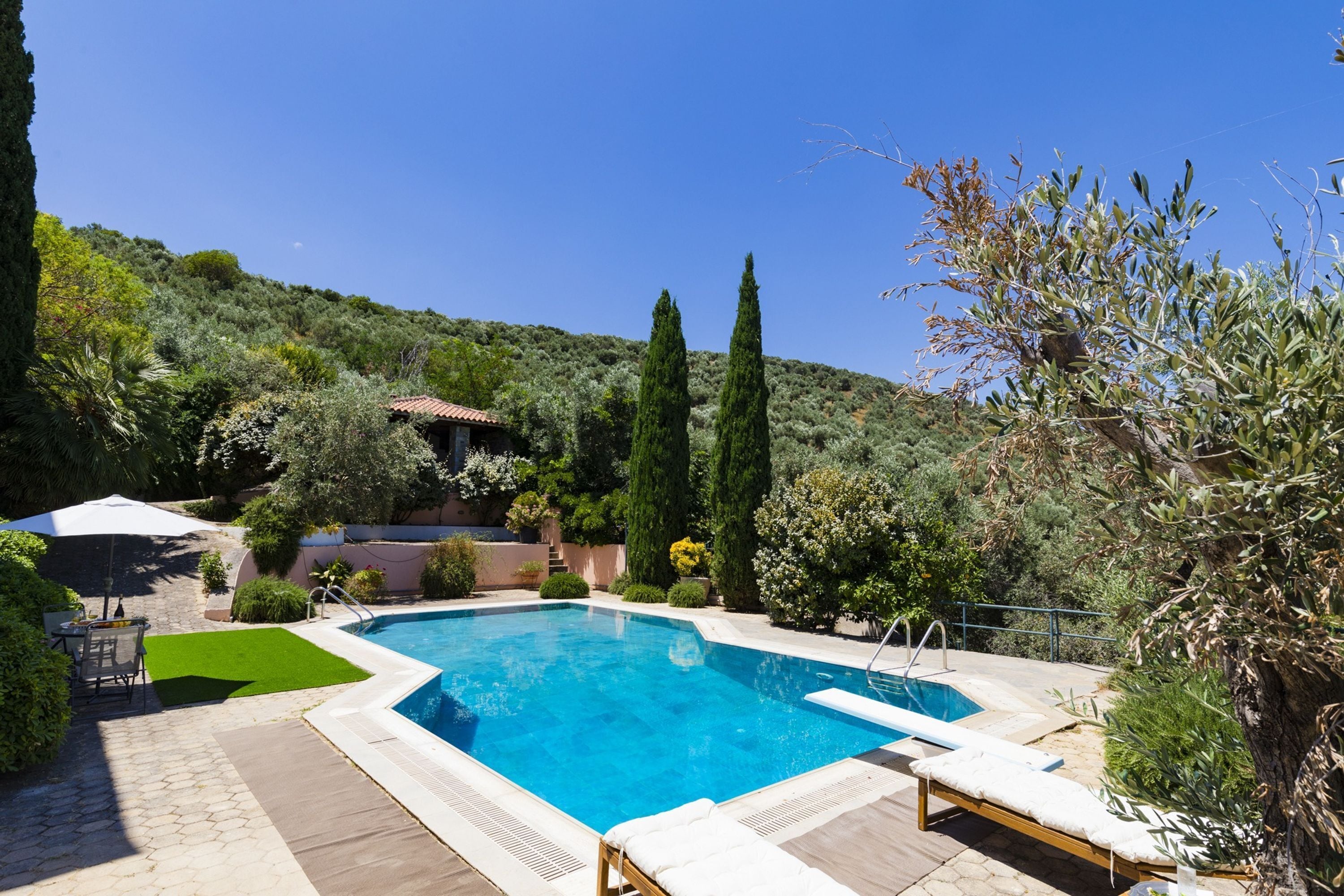 Une piscine rectangulaire, bordée d’une terrasse carrelée, entourée de verdure et située au pied d’une colline.