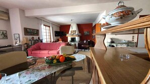 A living room with a pink sofa, a dining table with a floral tablecloth, and a kitchen area with a wooden countertop and a decorative teapot. - Alexia s Residence in Volos (Volos)