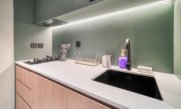 A modern kitchen with a stainless steel sink, a gas stove, a utensil holder, a soap dispenser, and a wall-mounted light. - Modern Studio Apt w Burj Khalifa View (Dubai)