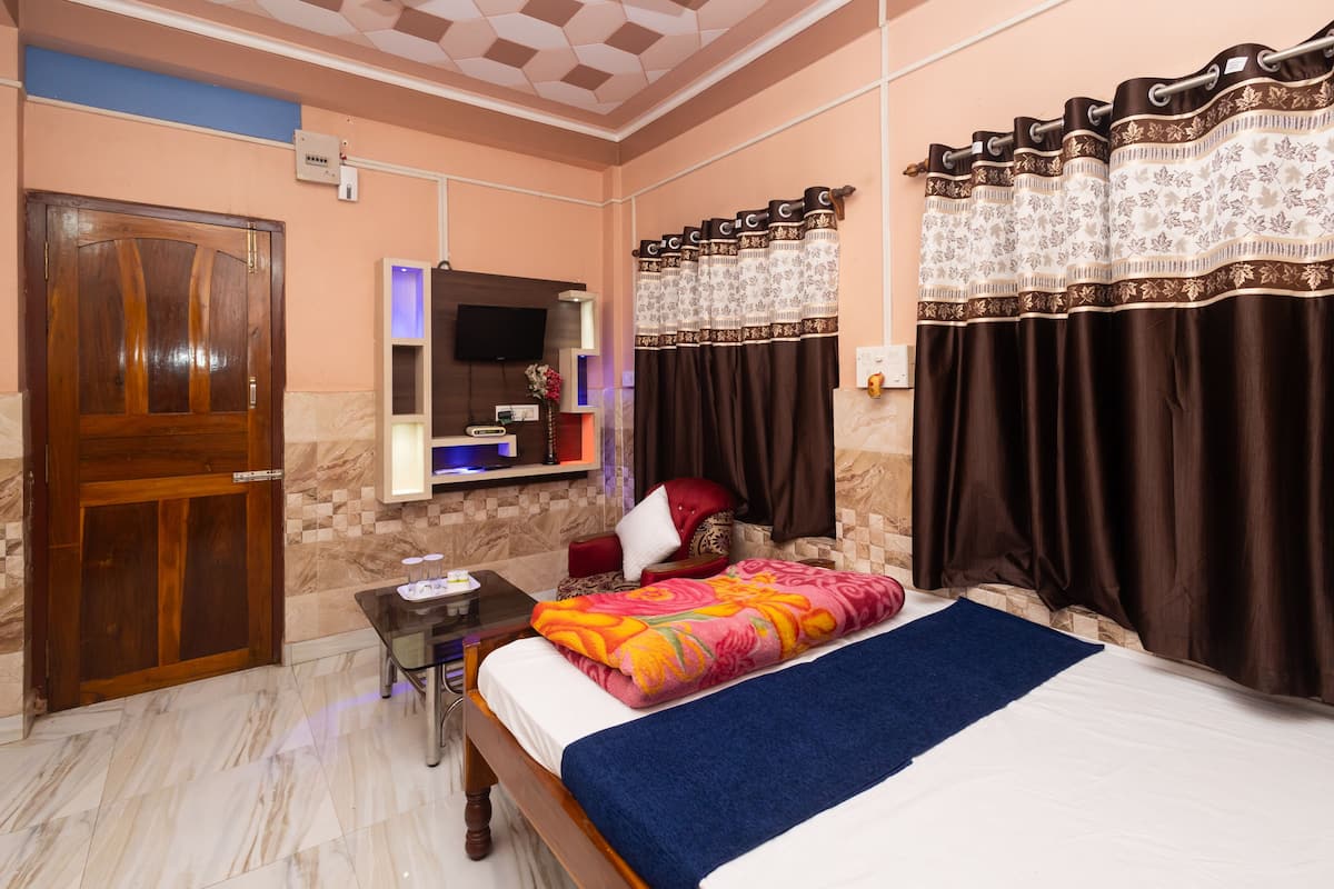 A hotel room with a wooden door, a bed with a colorful blanket, a small table with a lamp, a TV on the wall, and a bathroom with a shower curtain.