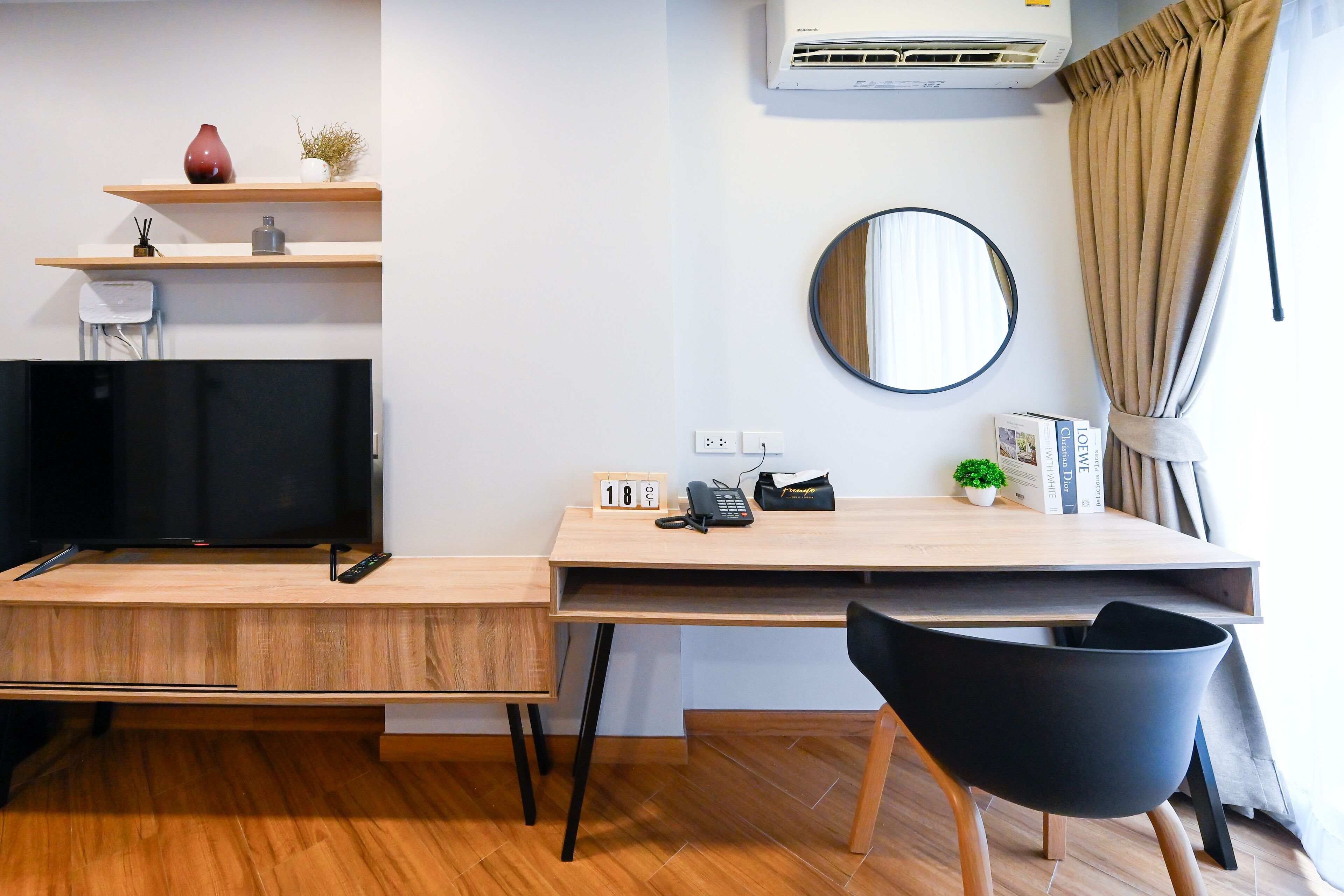 A modern room with a wooden desk, a black chair, a TV, a mirror, and a shelf with decorative items.