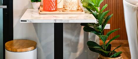 Una habitación moderna con una mesita de mármol, dos recipientes de vidrio con asas, una pequeña planta en maceta, una cesta de mimbre y un cubo de basura blanco.