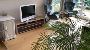 A living room with a TV on a wooden stand, a sofa, a chair, and a large window with curtains.