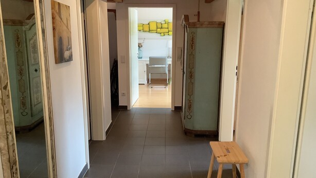A hallway with a wooden bench, a door with a decorative frame, and a view into a room with a kitchen area.