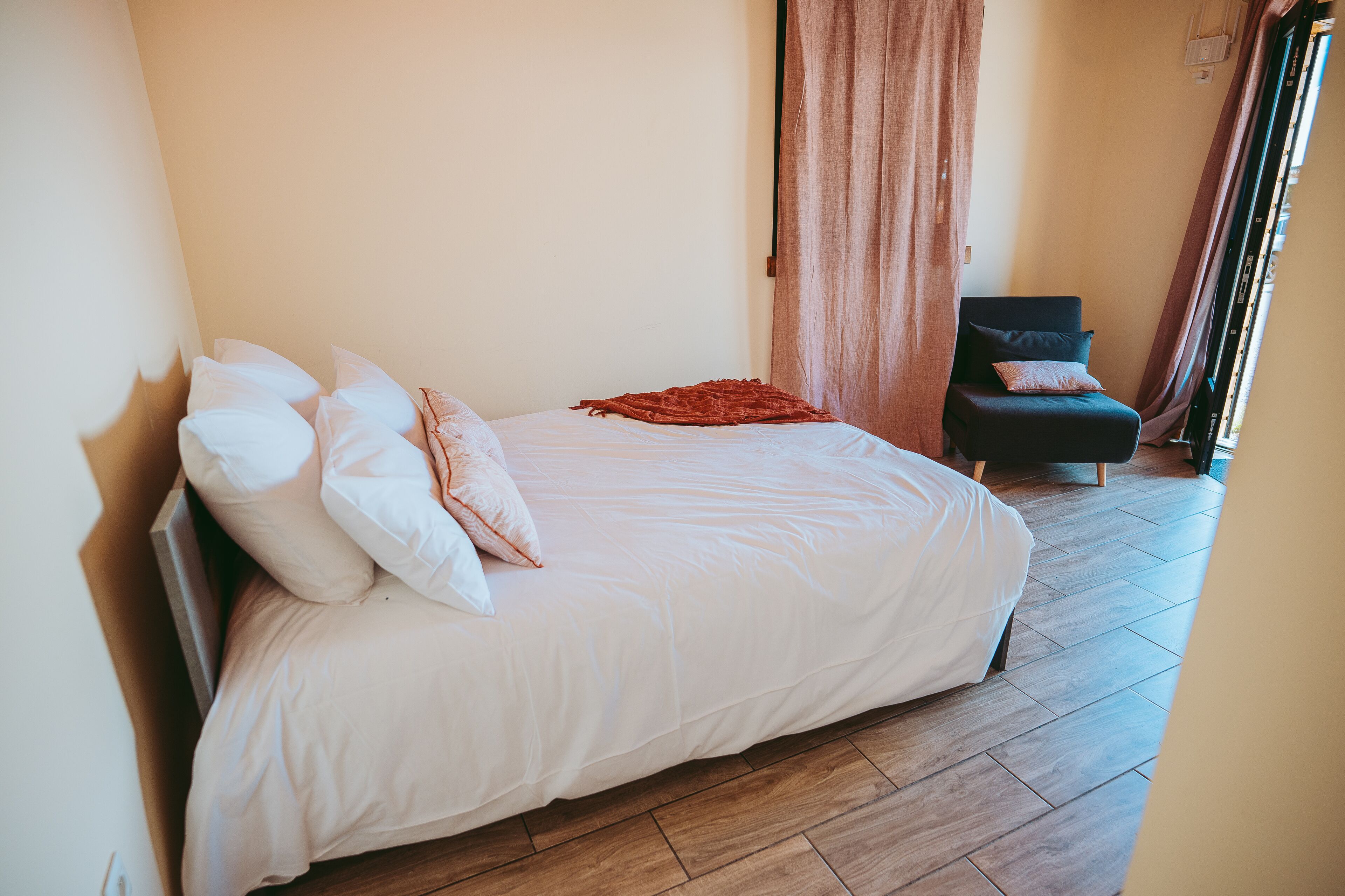 Ein ordentlich gemachtes Bett mit weißer Bettwäsche und mehreren Kissen, ein dunkler Sessel mit Sitzkissen und ein Holzboden.