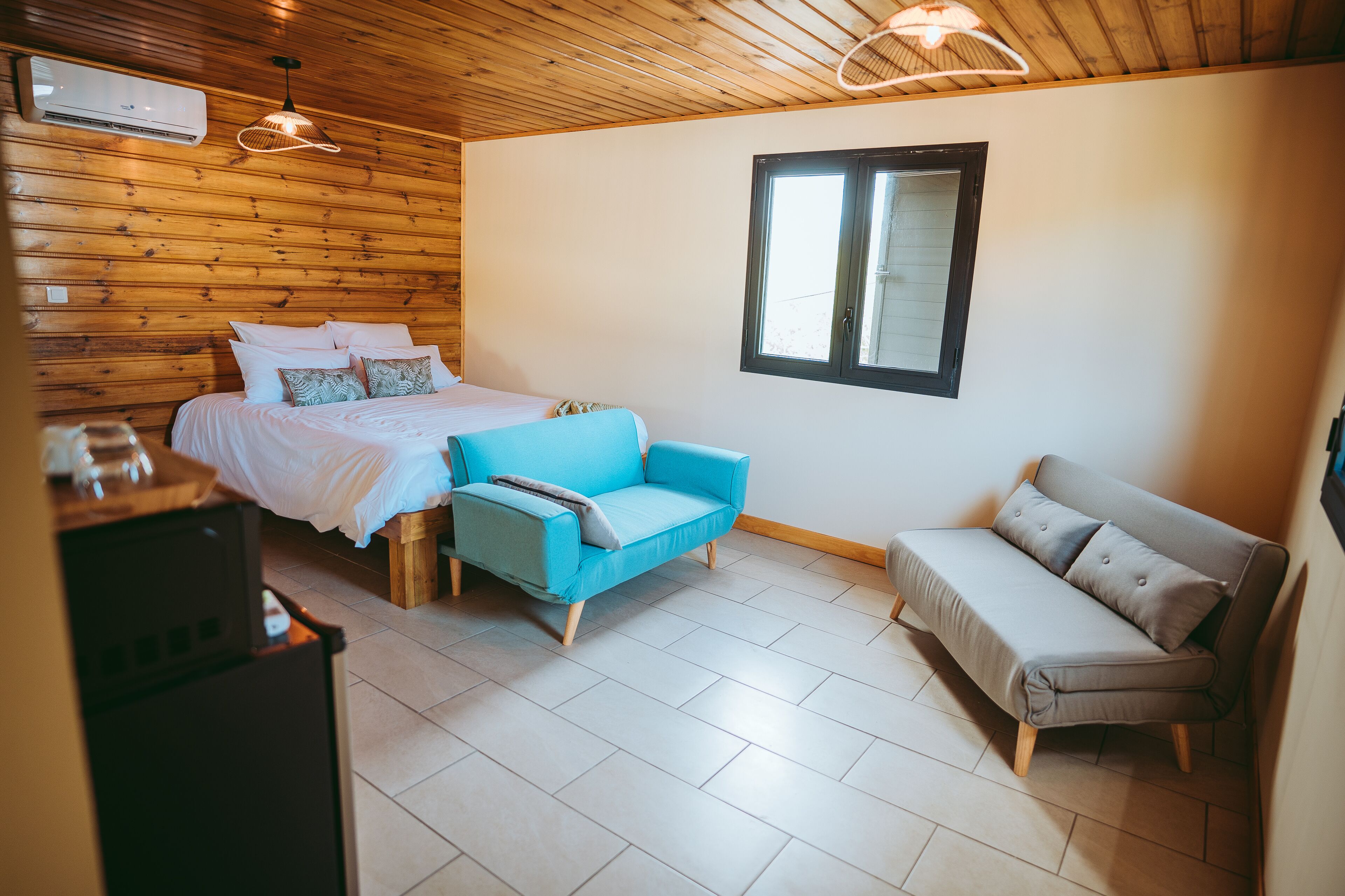 A room with a bed, a blue chair, a grey sofa, wooden walls, a window, and a ceiling fan.