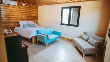 A room with a bed, a blue chair, a grey sofa, wooden walls, a window, and a ceiling fan.