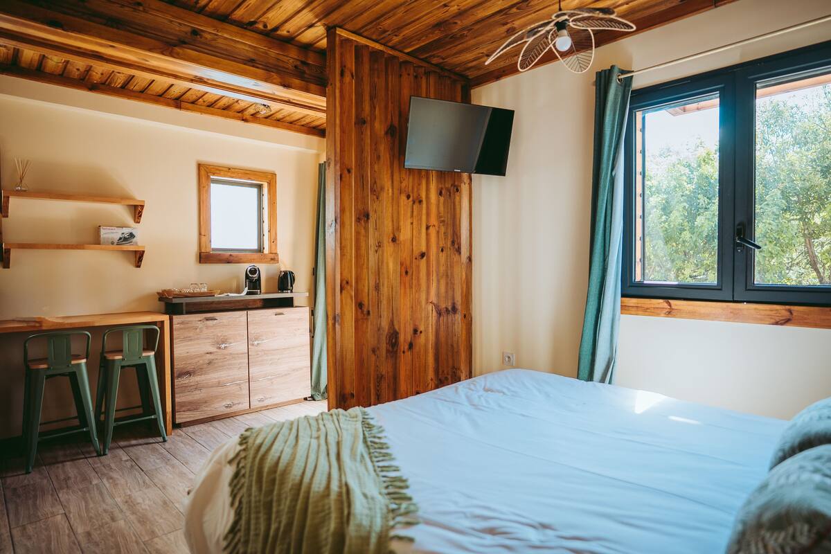 Une chambre avec un plafond en bois, un lit, un téléviseur fixé au mur, une fenêtre avec des rideaux et un bureau avec des chaises.