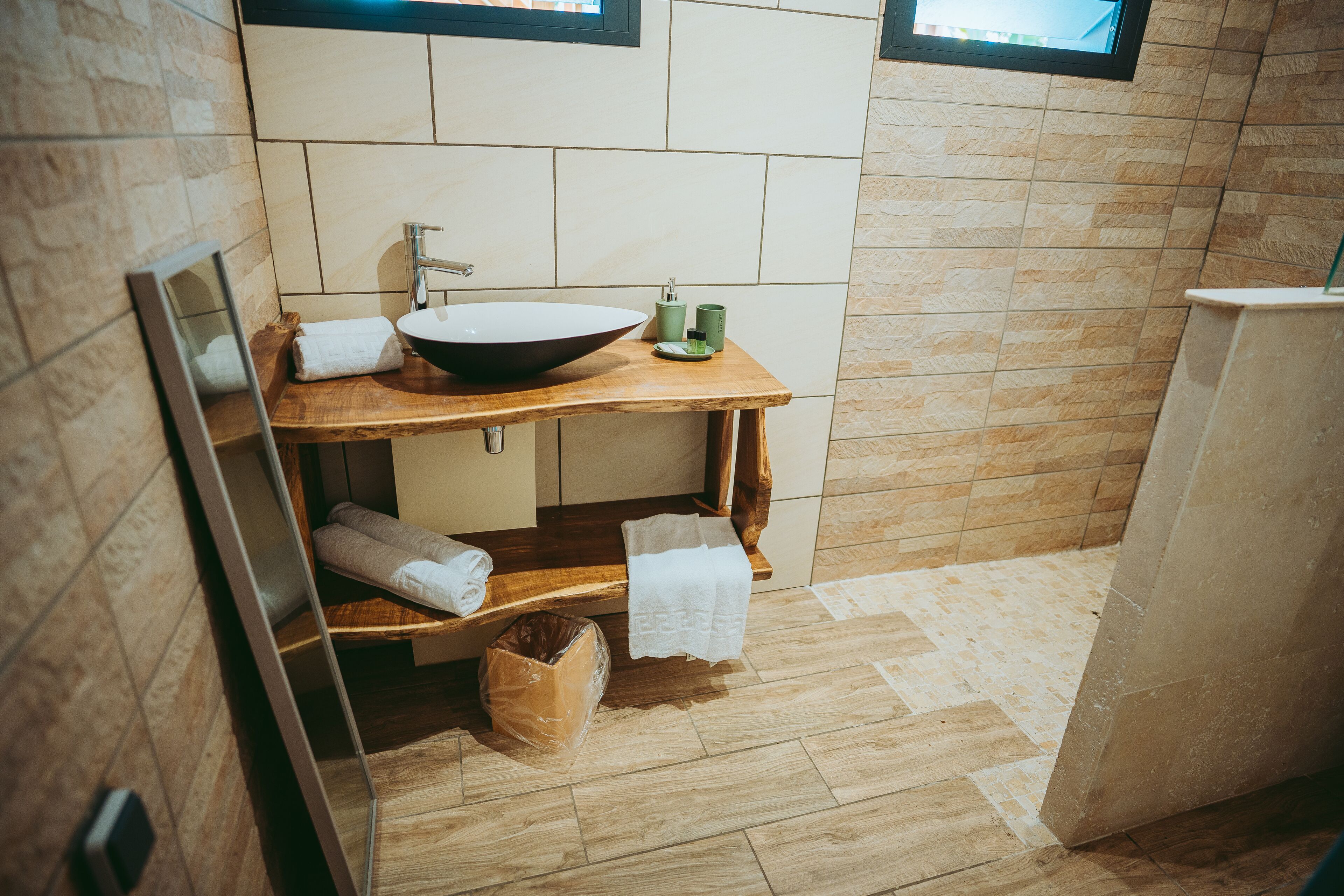 A bathroom with a wooden vanity, a large vessel sink, and a mirror. There are towels on the vanity and a trash bin on the floor.