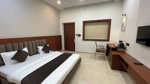 A hotel room with a large bed, a desk, a chair, a television, and a window with blinds. - The Kedar Resort (Mathura)