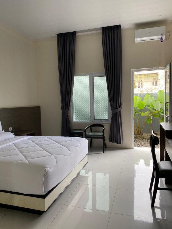 A hotel room with a large bed, a desk, two chairs, and a window with curtains. - Genio Syariah Hotel Solo (Surakarta)