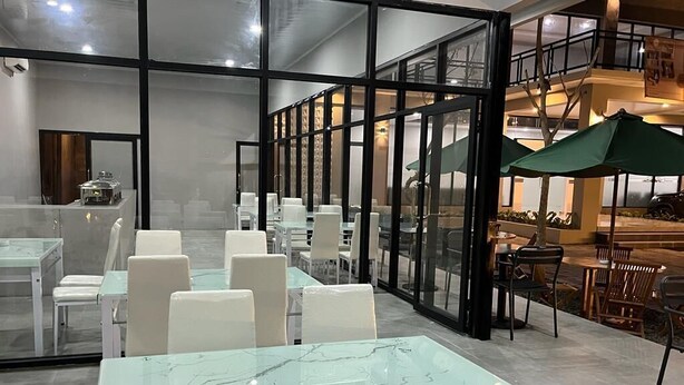 A modern indoor dining area with glass tables and white chairs, a glass partition, and a view of the outdoor seating area. - Genio Syariah Hotel Solo (Surakarta)