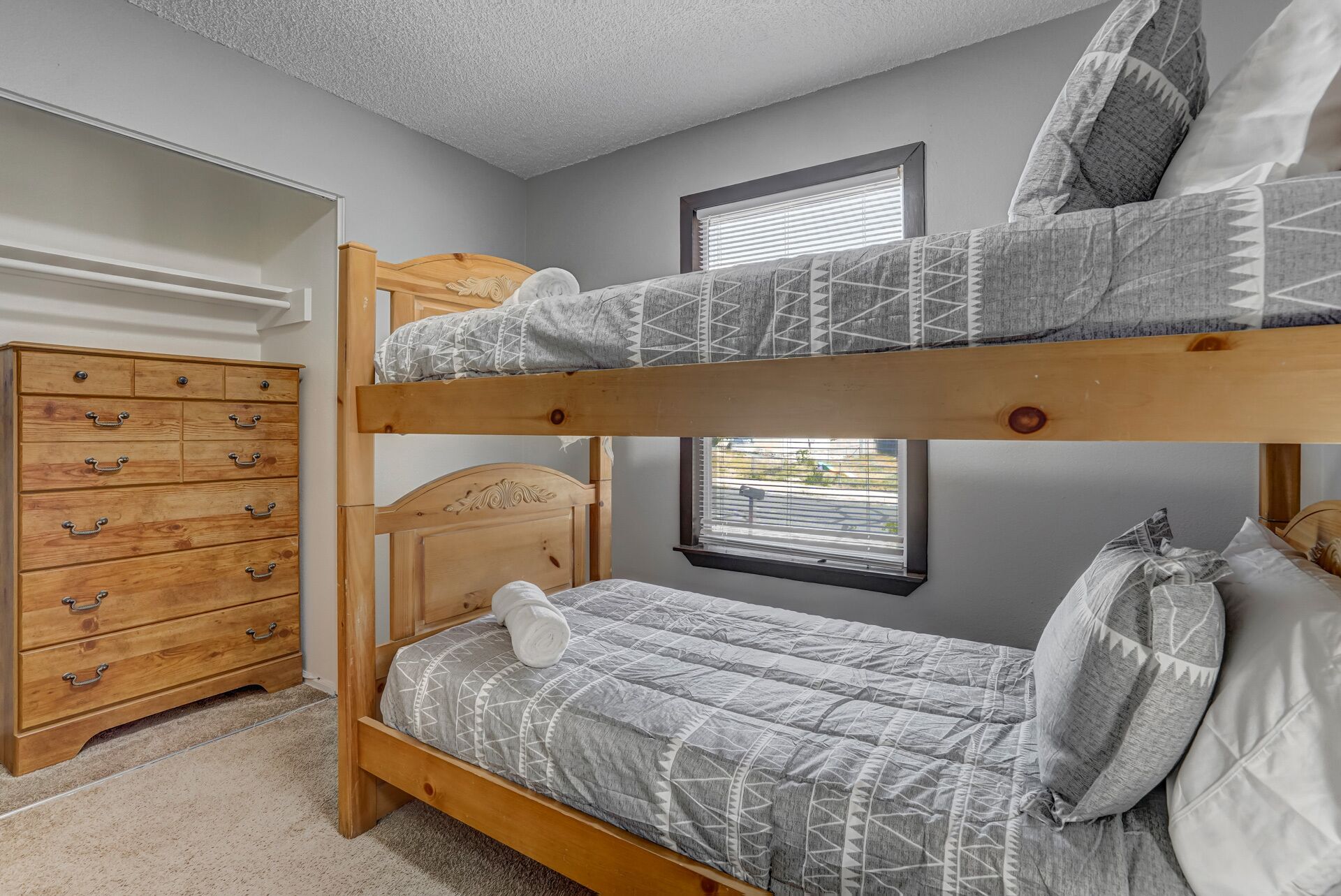 En våningssäng med ett mönstrat överkast, en byrå i trä med flera lådor och ett fönster med persienner.