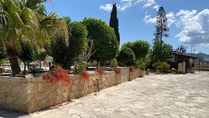 A paved courtyard with stone flooring, a stone wall with red flowering plants, and various trees including a tall conifer and a palm tree. - Beautiful, Unique, 'stone-built villa on the outskirts of Peyia  (Peyia)