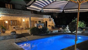 A swimming pool with a stone deck, a covered seating area with chairs and tables, and a stone building with a warm light. - Beautiful, Unique, 'stone-built villa on the outskirts of Peyia  (Peyia)
