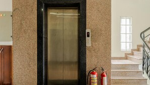 Un ascensor con pantalla digital, extintores y una escalera con barandilla metálica.