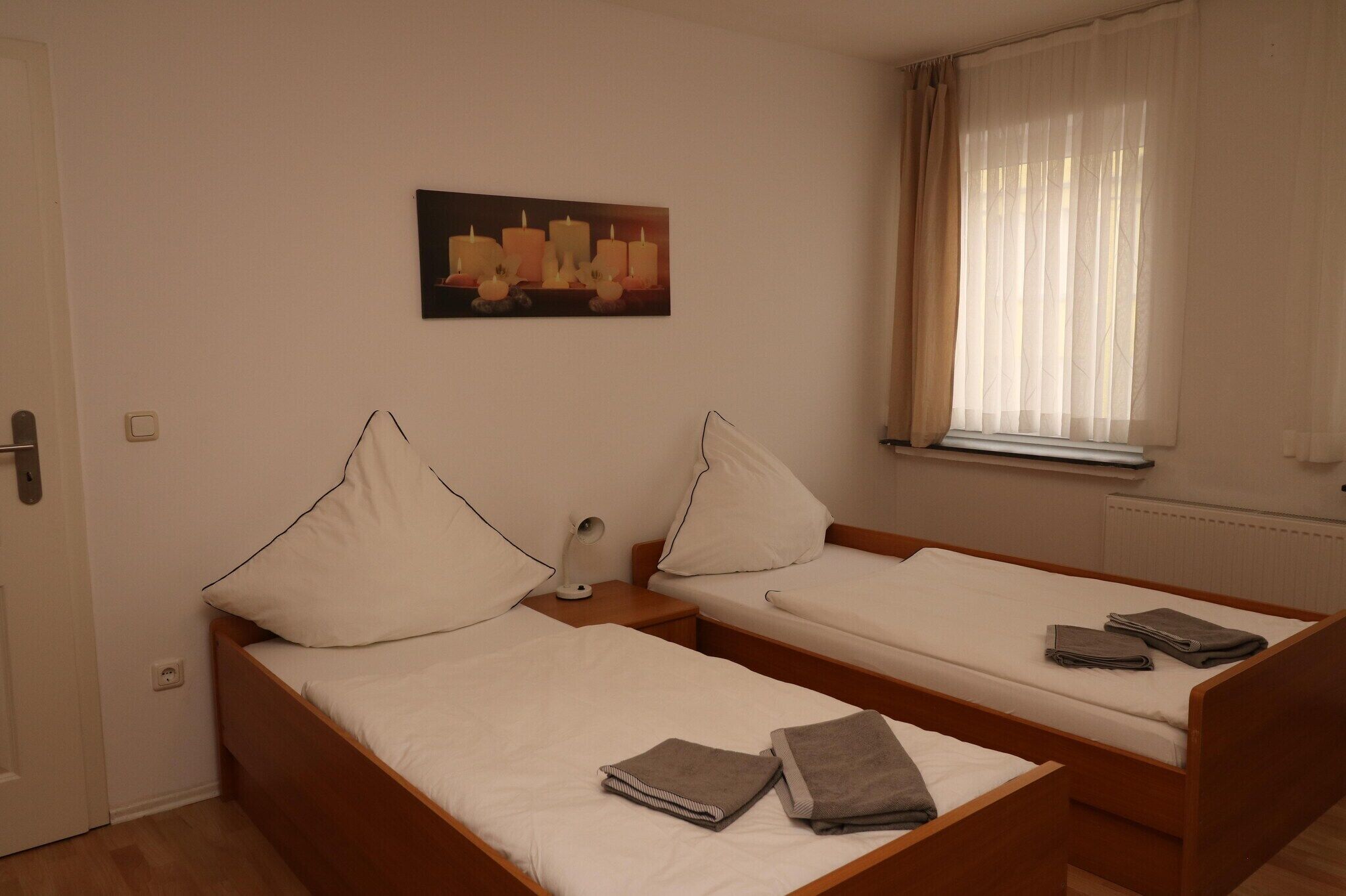 Quarto com duas camas de solteiro, mesa de cabeceira com abajur, um quadro na parede e janela com cortinas leves.