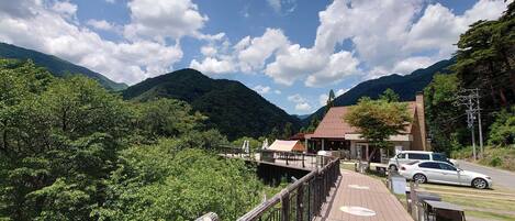 A wooden deck with a railing, leading to a building with a red roof and brick walls, surrounded by lush greenery and mountains.