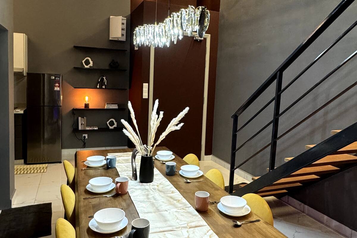 A modern dining area with a wooden table set for six, surrounded by yellow chairs. A chandelier hangs above the table, and a staircase is visible to the right.