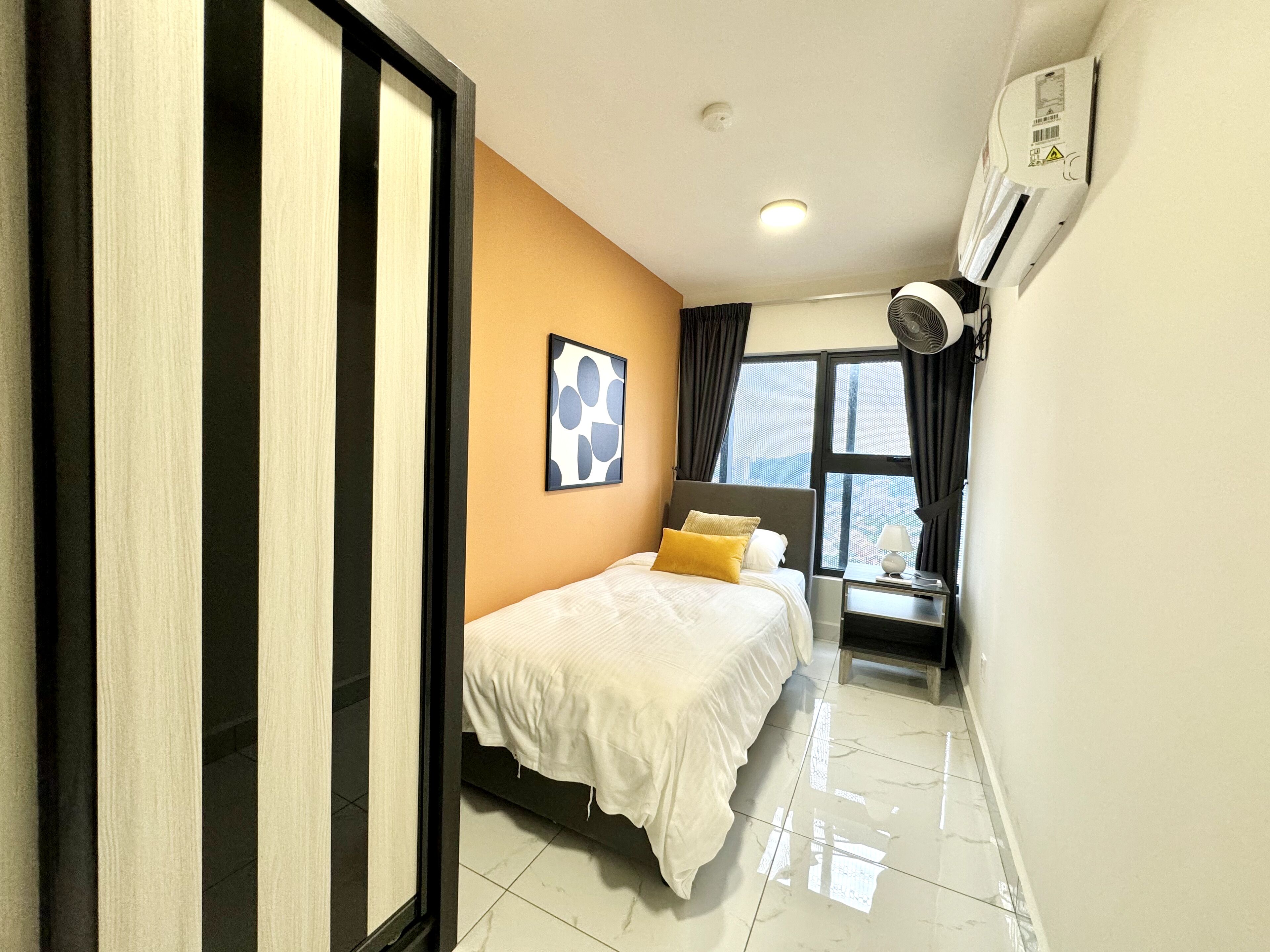 A hotel room with a bed, a bedside table with a lamp, a window with curtains, and an air conditioning unit on the wall.