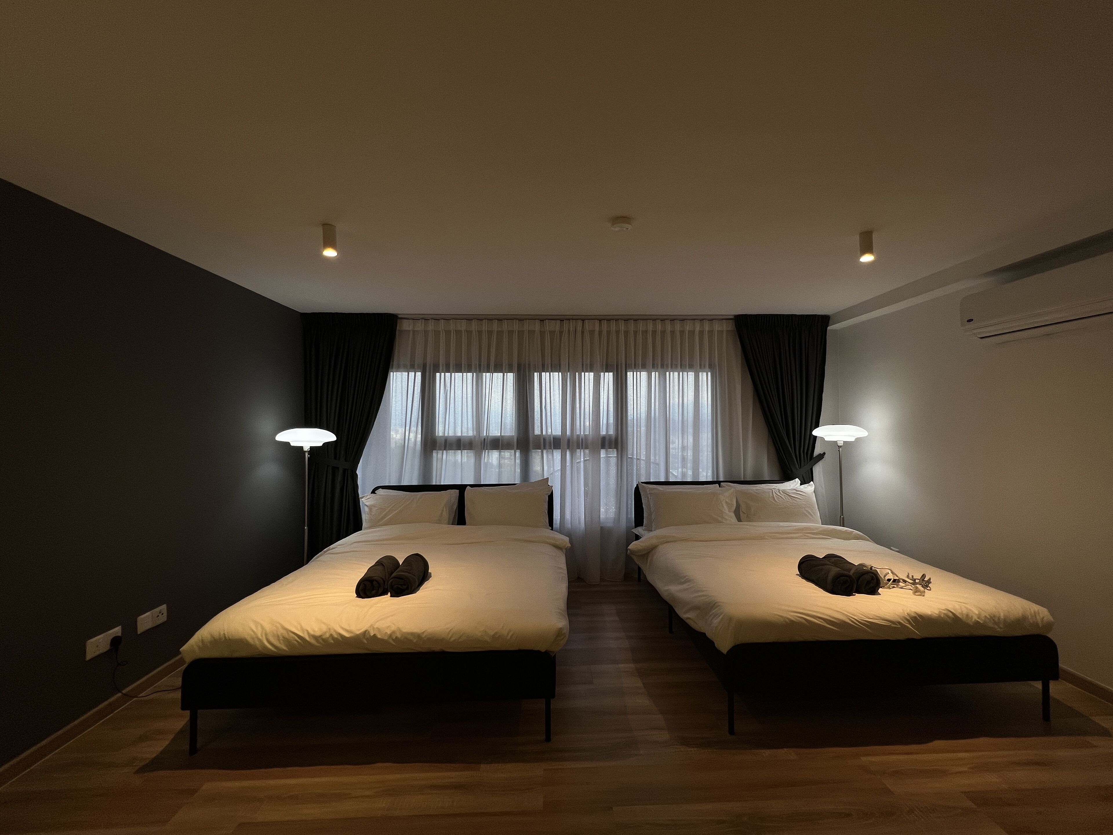 A hotel room with two beds, a large window with sheer curtains, and two bedside lamps.