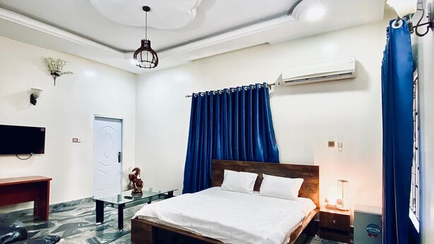 A bedroom with a bed, a nightstand, a desk, a chair, a wardrobe, and an air conditioning unit. - Hilton Summer Apartments (Abuja)