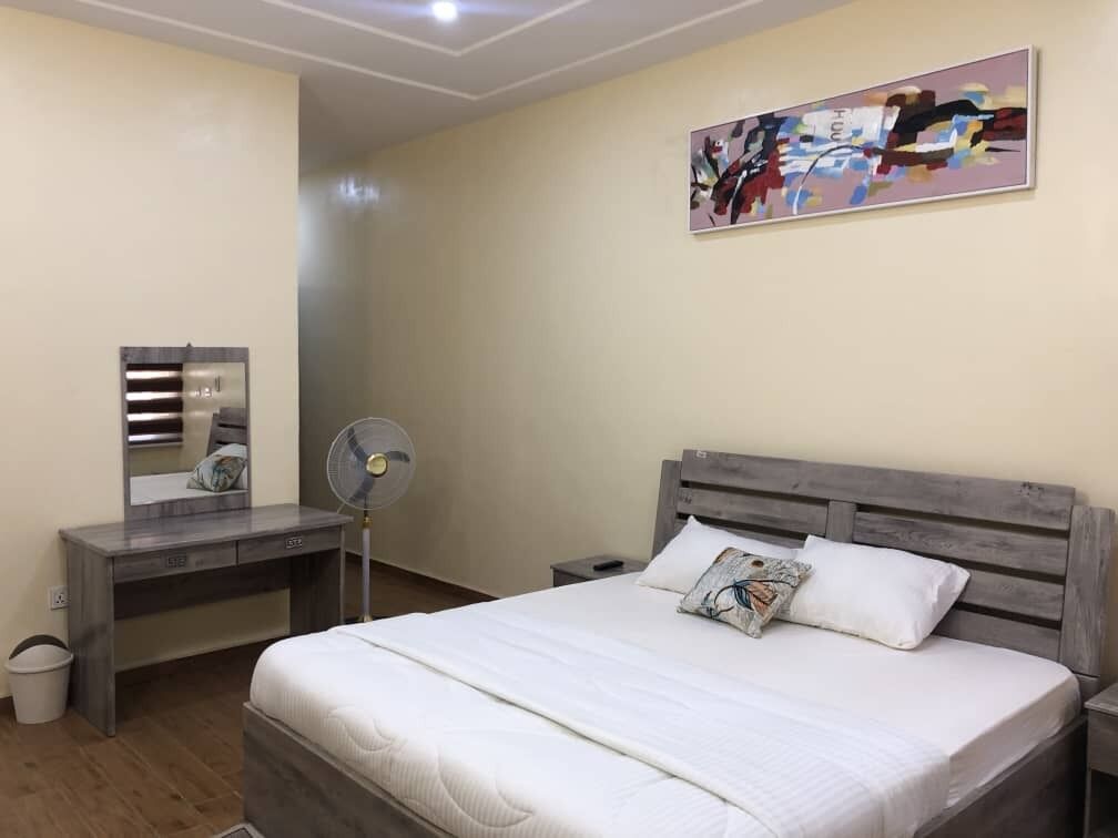A bedroom with a bed, a desk, a chair, a fan, and a wall-mounted artwork.