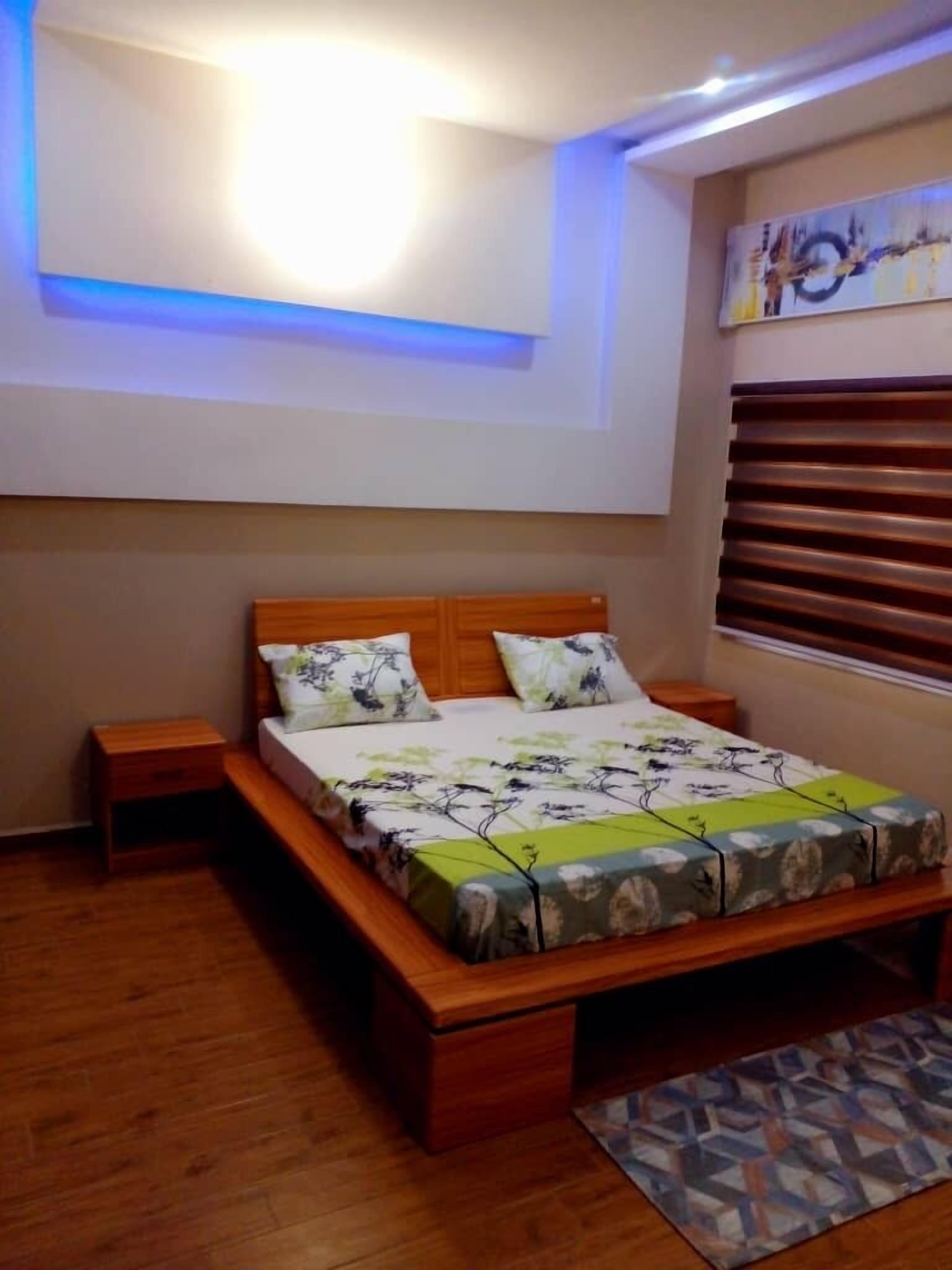 Un dormitorio con una cama de madera, una mesita de noche, una ventana con persianas y una lámpara en el techo.