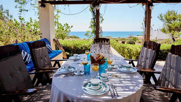 An outdoor dining area with a view of the sea, covered with a wooden pergola, and furnished with striped chairs and a round table set for a meal.