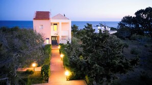 A two-story building with a red-tiled roof, a pathway leading to the entrance, and a view of the sea in the background.